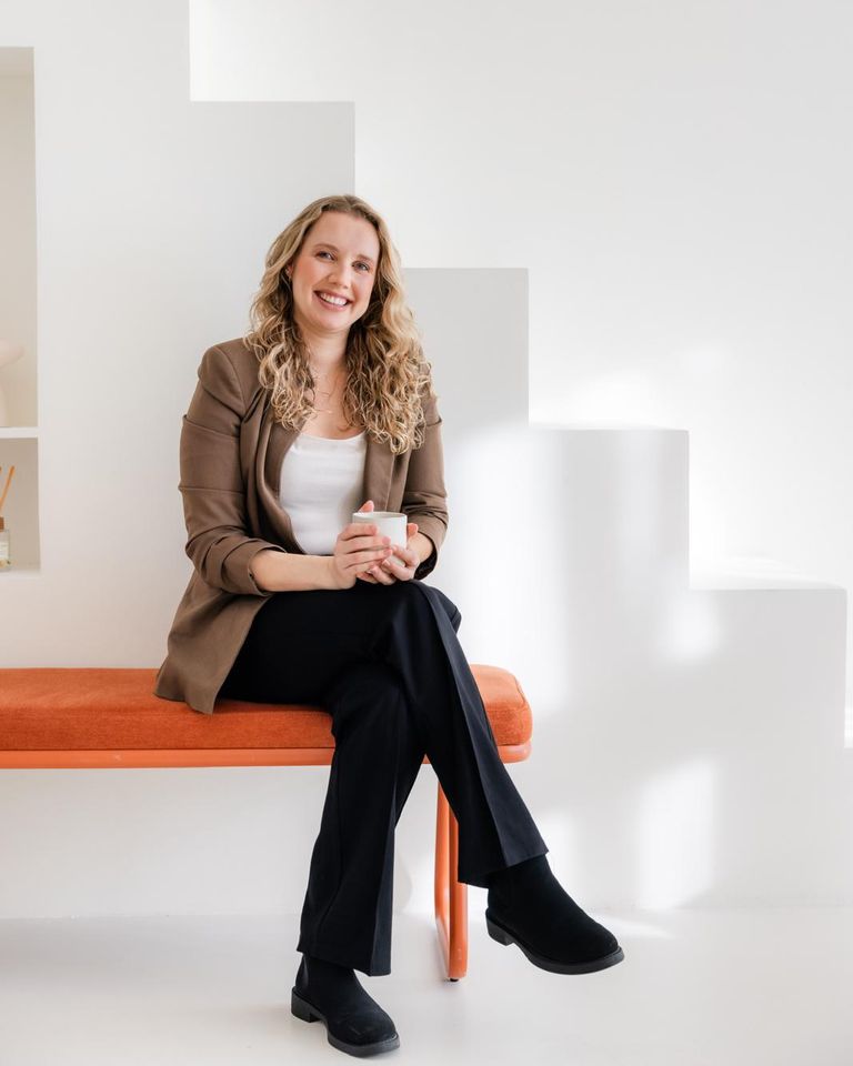 Debra Bekker zit lachend op een oranje bank in een lichte hal